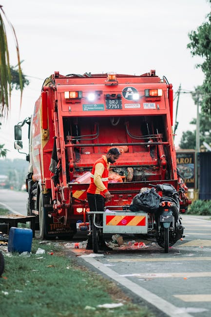 A sanitation worker wearing a yellow and orange high-visibility vest is using a manual tool to collect waste from a black plastic bag placed on the ground near the rear of a red trash collection vehic