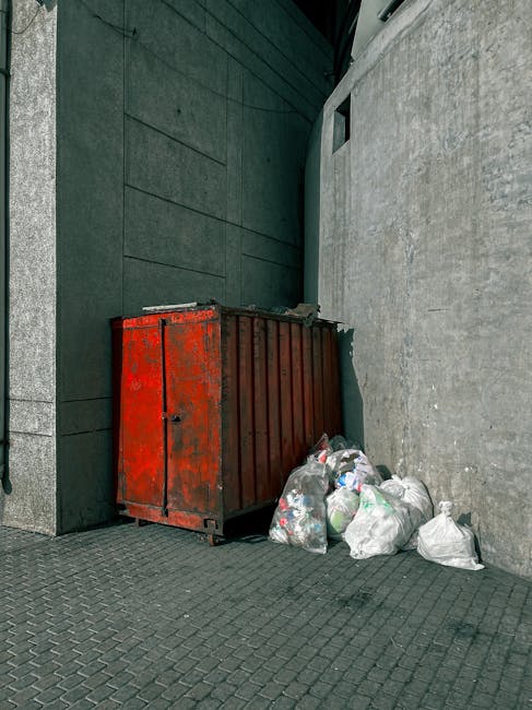 A sanitation worker wearing a yellow and orange high-visibility vest is using a manual tool to collect waste from a black plastic bag placed on the ground near the rear of a red trash collection vehic