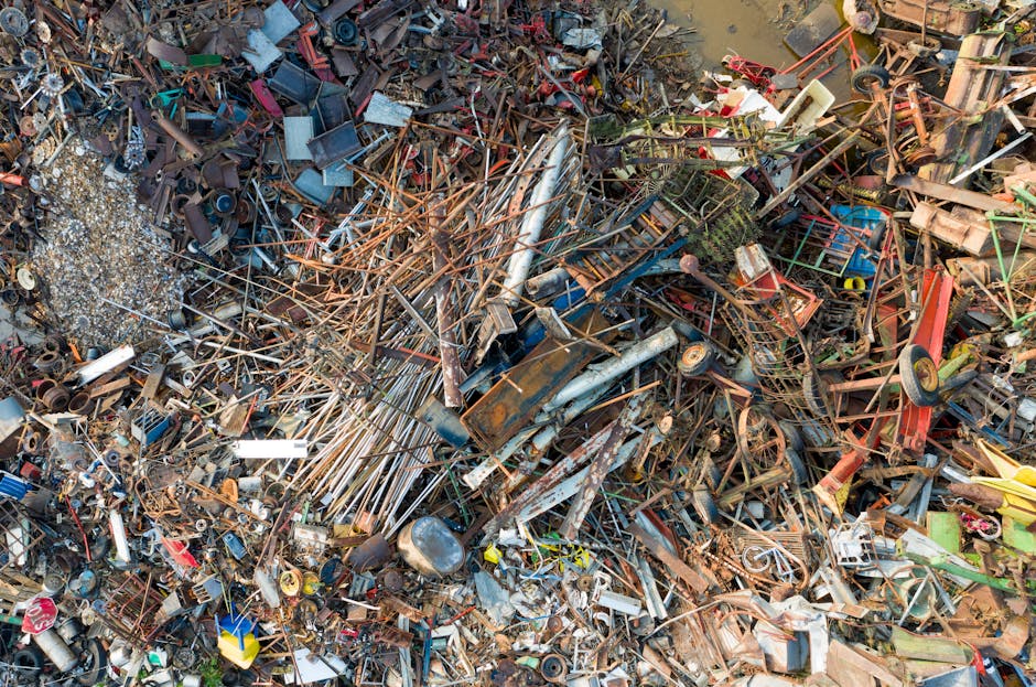 An aerial view of a large, unorganized landfill site filled with a mixture of waste materials including plastic bottles, packaging, textiles, and miscellaneous debris. The waste is scattered across a 