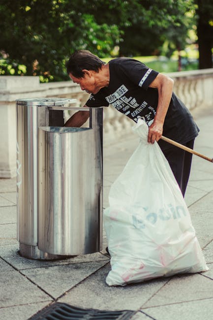 A clear cylindrical waste bin made of transparent plastic material is positioned on a beige carpeted floor, with crumpled white paper scattered around it. The crumpled papers vary in size and shape, s