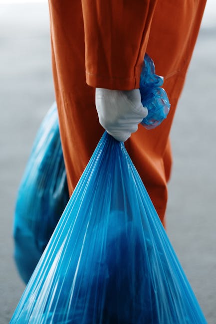 The image shows a person dressed in orange work overalls standing on a smooth, grey concrete floor against a plain light grey background. They are holding two large, semi-transparent blue plastic rubb