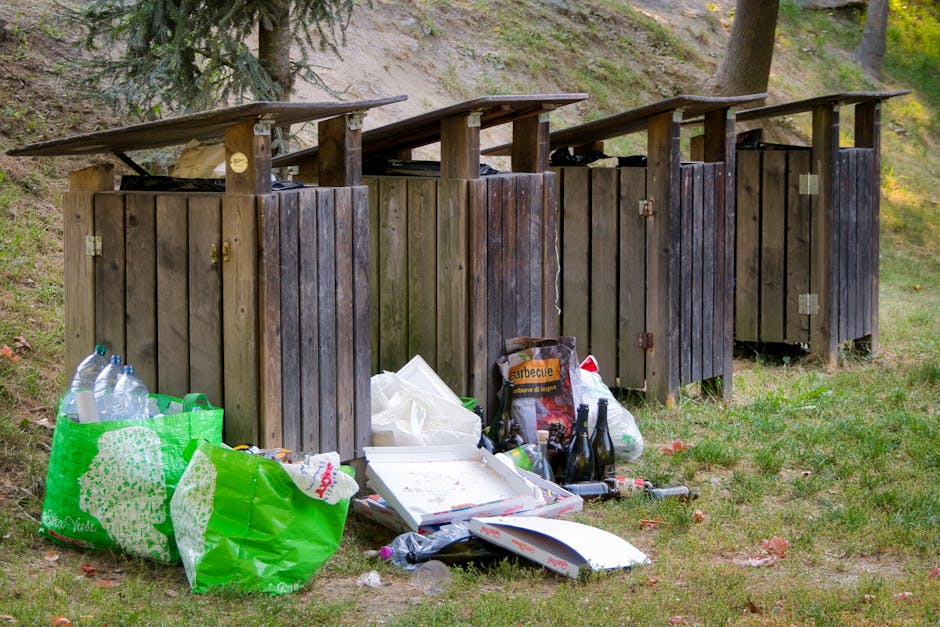 A row of four wooden outdoor rubbish bins with hinged lids, set on a grassy area near a sloped, wooded background. The bins are constructed from vertical wooden planks with visible metal hinges and ha